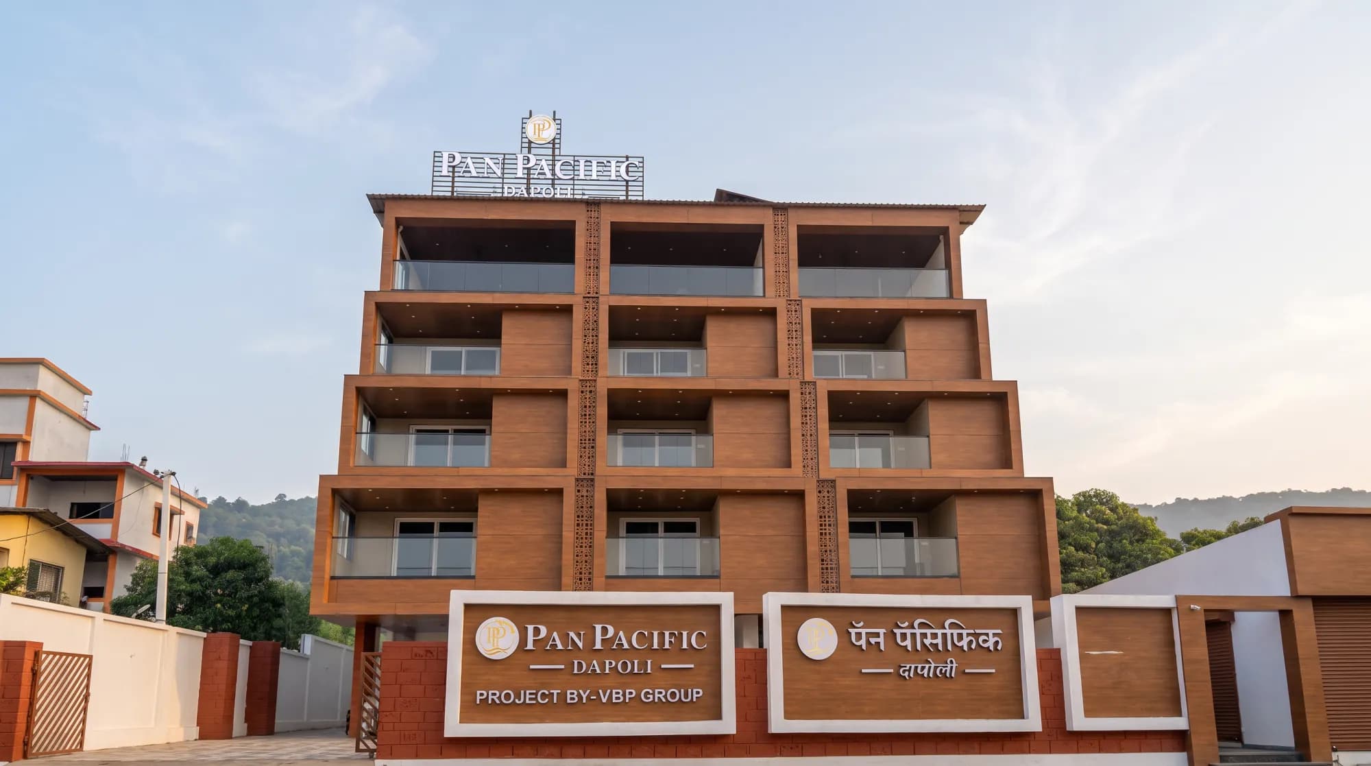 The terracotta façade of Pan Pacific Dapoli with its signboards, against green hills