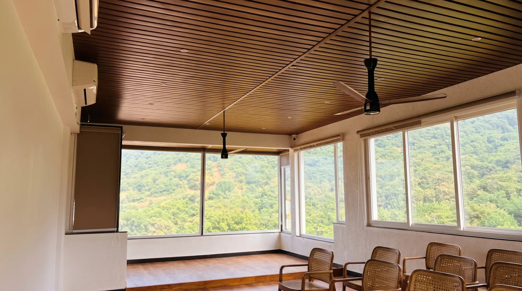 Cane chairs beneath a wooden slatted ceiling, with wide windows onto wooded hills